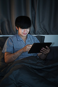 坐在床上 晚上用数字桌子微笑着的亚洲男人 (笑声)图片