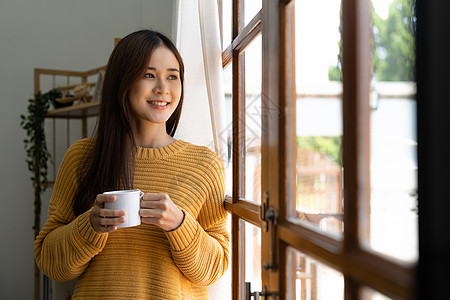 快乐的亚洲女人在窗户旁边喝着热咖啡 笑着带着舒适的家图片