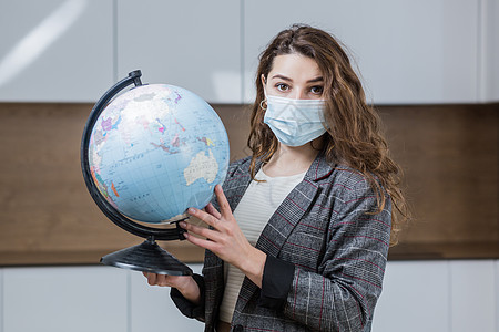 一位戴着医用口罩远程教授在线地理课的年轻女教师的肖像 她拿着一个地球仪 指着一个全球地球仪 看着相机图片
