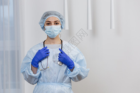 戴面罩 外科长袍和帽子的年轻女医生肖像 站立拿着听诊器 看镜头图片