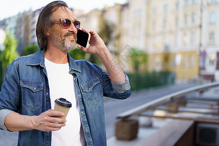 英俊的灰胡子中年男子在电话里聊天 手里拿着一次性纸杯里的咖啡 站在户外 穿着牛仔裤衬衫 自由职业者旅行人背景图片