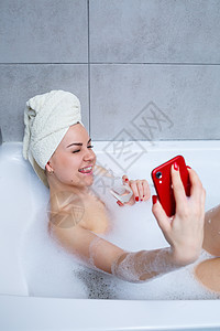 头上裹着毛巾的女孩躺在白色浴缸里 手里拿着一杯香槟 正在打电话自拍 她手里拿着很多醉肥皂 辛苦一天后放松一下 水疗放松程序享受福图片