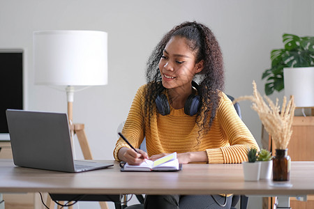 微笑的年轻非洲裔美国女性戴着耳机 从电脑工作中分心 看着远方的梦 快乐的混血儿女性在家里通过笔记本电脑思考或计划在线学习 视觉概图片