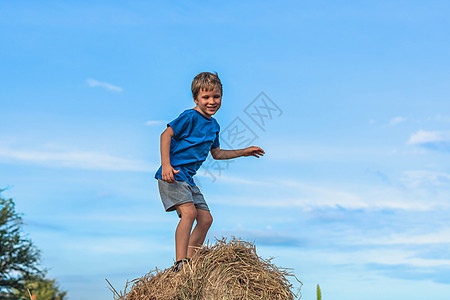 男孩微笑着玩着舞蹈鬼脸炫耀蓝色 T 恤站在干草堆上 晴朗的天空阳光灿烂的日子 平衡训练 快乐童年的概念 户外儿童 接近大自然的清图片
