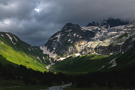 充满鲜青的绿河谷 河流在草地上 树木在雪积岩石山脚下 有瀑布 大片闪亮的雷声云 阳光穿透爬坡旅行山脉顶峰岩石森林峡谷场景远足全景图片