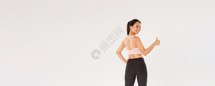 全长充满动力和满意的微笑亚洲女教练 身穿运动服的女运动员转身对着镜头竖起大拇指 准备进行富有成效的健身训练 白色背景工作室运动装图片