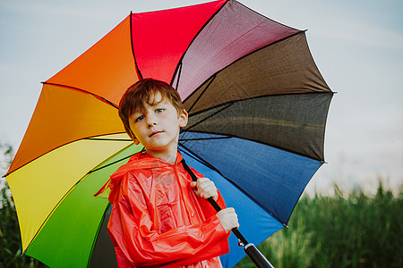 公园里拿着彩虹伞微笑的男生画像 孩子在他的肩上拿着五颜六色的雨伞 穿着红色雨衣的快乐孩子在户外拿着多色雨伞池塘橙子彩虹绿色学校自图片
