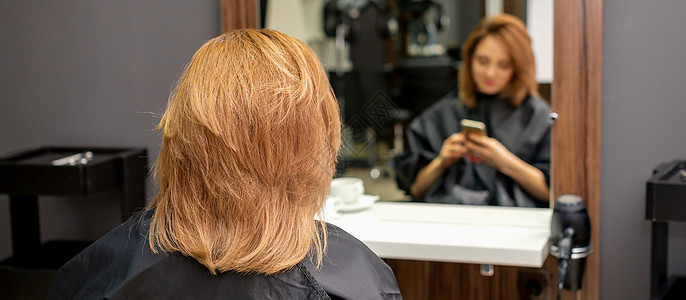 美丽的年轻红发女郎用她的智能手机 在镜子前发短信 等着看理发师的戏呢 笑声成人互联网工具电话女性客户技术护理头发奢华图片