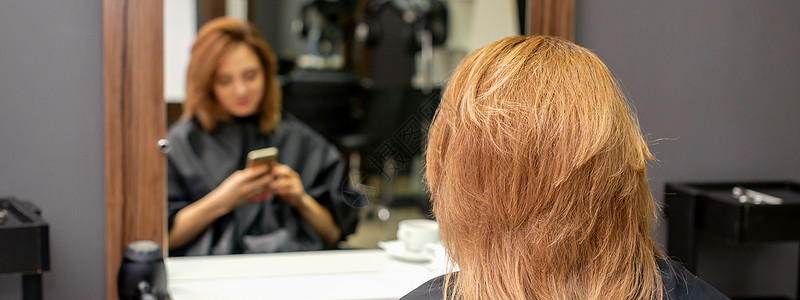 美丽的年轻红发女郎用她的智能手机 在镜子前发短信 等着看理发师的戏呢 笑声治疗理发反射女性顾客细胞成人优雅头发发型图片