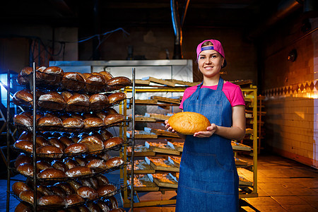 专业面包师  一位身穿牛仔裤围裙的年轻漂亮女性在面包店或面包店的背景下拿着新鲜面包 烘焙食品 面包生产职业购物职场食物工厂孩子工图片