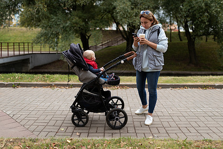 一位年轻母亲在城市周围的婴儿车里与婴儿一起散步时喝咖啡和看手机 并随婴儿行走图片