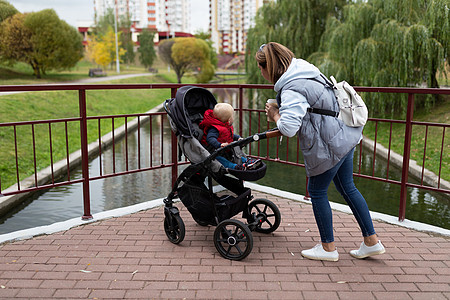 一名年轻母亲肩上背着背包 带着孩子在市公园的婴儿园里散步图片
