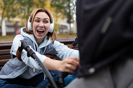 坐在长椅上的年轻母亲户外听耳机播放音乐 在婴儿步车中与婴儿玩感情游戏图片