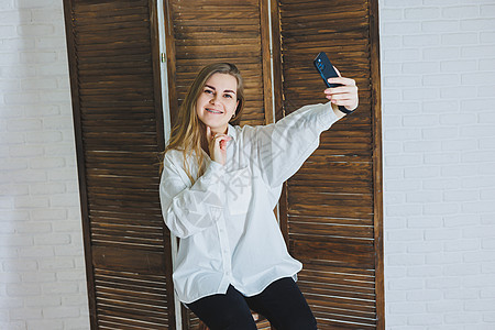身穿白色棉质衬衫 情绪激动的年轻女性指着手势 看着现代小工具的屏幕 一个拿着手机的女孩自拍博主摄影青年电话成人青少年派对照片互联图片