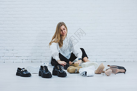 身着白色衬衫和黑色裤子的时髦服装 微笑着的快乐美丽的年轻女人坐在鞋子中间的地板上 为自己选择一双新鞋脚跟女性衣服房间衣柜地面女孩图片