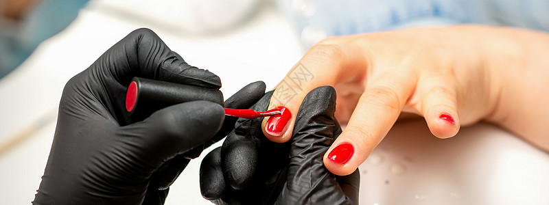 在美容院的一只雌性指甲上涂着红色清漆 穿着橡皮黑手套的美甲师傅贴近了她的身影绘画一部分护理服务化妆品抛光横幅手指奢华女士图片