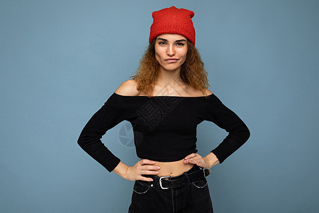 身着黑作物顶顶和红帽子的蓝色背景墙上被孤立 看着摄影机 校对红帽帽子黑色裁剪上衣女性自信卷曲图片
