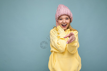 照片中 美丽 微笑的年轻金发女子被隔离在蓝色背景墙上 戴着冬季粉色帽子和黄色连帽衫 看着相机 指着你图片