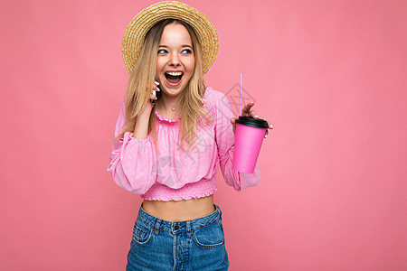 照片中 美丽积极情绪化的年轻金发女性身穿粉色作物衬衫和草帽 一边喝着粉色背景中带有复制空间的饮料 一边用手机聊天成人金发女郎咖啡图片