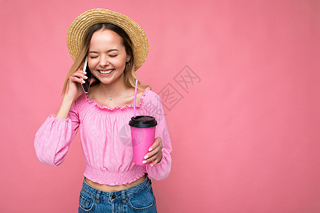 快乐的年轻女子在用手机聊天时 在喝粉红背景咖啡时交谈金发女郎女性情绪情绪化衬衫幸福空间纸杯作物饮料图片