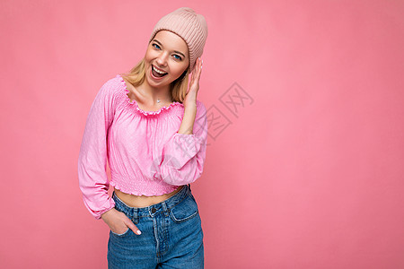 美丽 积极 快乐的金发女郎 与色彩缤纷的背景墙隔开 穿着休闲时尚的衣服 看着镜头 心情真挚裁剪金发青年潮人粉红色空间粉色衬衫帽子图片