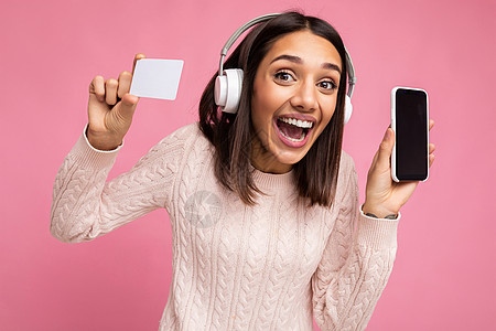 迷人 积极 快乐 微笑的年轻女性穿着时尚的休闲装 戴着白色无线耳机 与彩色背景墙上隔开 拿着塑料信用卡 展示手机 屏幕空空 可以图片