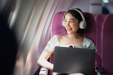 在飞行 旅行和商务概念期间 亚洲青年女子在飞机头等舱靠近窗户窗口的飞机上使用笔记本电脑戴耳机男性喷射人士班级女士工作女孩空气运输图片