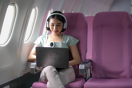 在飞行 旅行和商务概念期间 亚洲青年女子在飞机头等舱靠近窗户窗口的飞机上使用笔记本电脑戴耳机人士空气技术女士女性乘客班级女孩男人图片