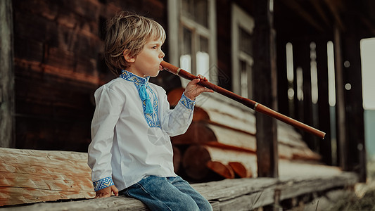 小男孩演奏木管乐器木笛-乌克兰 sopilka 民间音乐概念 乐器 穿着传统刺绣衬衫的孩子 坐在老木屋门廊上的孩子图片