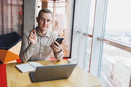 成功的年轻人正坐在室内一家咖啡馆餐厅的桌子旁 在笔记本电脑上工作和学习 手里拿着一部空白屏幕的手机 自由职业者移动办公室经营理念图片