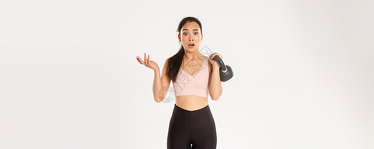 运动 健康和积极的生活方式理念 困惑的亚洲健身女孩 女运动员举起壶铃 看起来很困惑 在训练期间咨询教练 站在白色背景身体广告女性图片