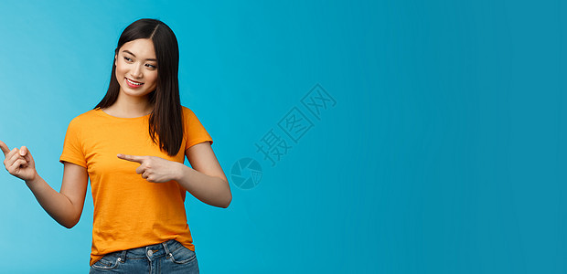温柔可爱的亚洲女孩 留着深色头发 好奇地指着左边 对店里的新产品感兴趣 站在蓝色背景上逗乐 娱乐 观看展览 穿黄色 T 恤皮肤女图片