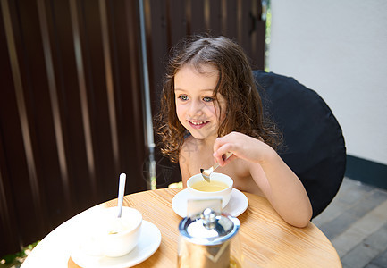 可爱的高加索小女孩坐在城市夏季露台的桌子上 喝茶 笑着甜美的笑容微笑图片