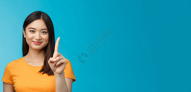 特写开朗 迷人 柔嫩的亚洲女性 留着深色短发 展示第一名 伸出食指 快乐地微笑 友好的笑容 站在蓝色背景放松 随便下单护理广告商图片