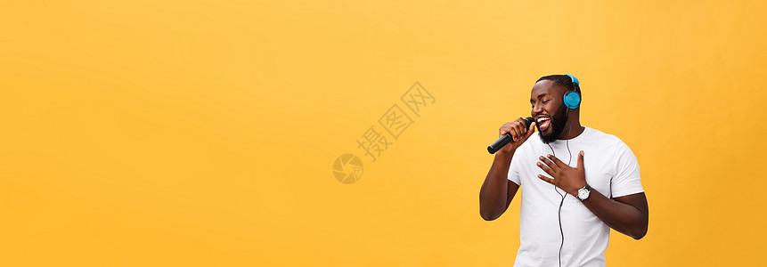 欢快积极 别致 英俊的非洲男子手持麦克风 头戴耳机听音乐唱歌 享受黄色背景中孤立的周末假期的肖像女孩青年寒意乐趣电话打碟机女性音图片