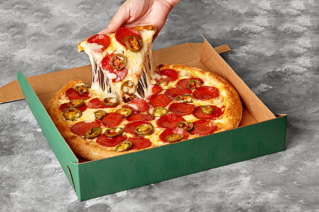 女性用手从纸板盒中拿一块披萨 加上融化的奶酪 辣椒尼和黄青椒图片