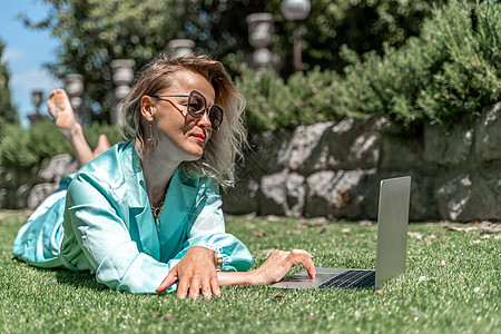 一位美丽的年轻女子 金发卷发戴眼镜 穿着蓝裙子 在大自然的草地上坐着 用笔记本电脑药片咖啡旅游学生金发女郎沙发房间蓝色女孩稻草图片