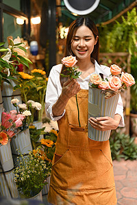 正面的年轻女小企业家穿着围裙 手捧粉红玫瑰花站在花店前图片