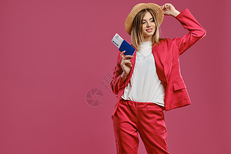 穿着草帽 白上衣和红色短裤的金发女性 微笑 持有护照和票 同时假扮粉红工作室背景图片