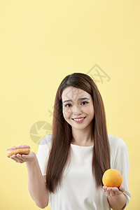 一个微笑的年轻亚洲女性 选择甜甜圈和橙色之间 孤立在黄色背景之上的肖像饮食食物女士水果营养白色图片