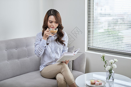 有吸引力的年轻女子阅读书本和喝咖啡 坐在家窗户附近的咖啡女性长椅沙发女孩房间图片