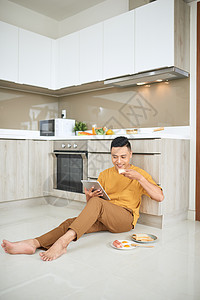 亚洲年轻人在家里工作 拥有网络生意 吃香肠和看平板电脑 笑声谷物厨房享受技术食物早餐触摸屏药片杯子男性图片