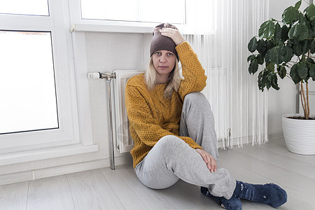 一个戴着帽子和黄色毛衣的年轻女孩坐在地板上 头靠着一个加热器用自动调温器图片