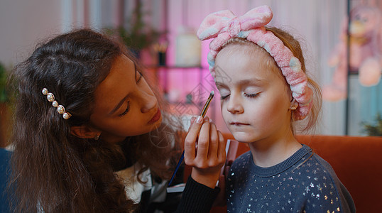 十几岁的孩子在家里的游戏室里帮她的小妹妹做面部化妆绘画兄弟姐妹沙发导师皮肤长椅护理朋友们口红友谊图片