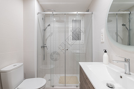 宽敞的白色瓷砖浴室被明亮的白色自然光照亮 墙上的大圆镜映照出带玻璃栏杆和金属配件的淋浴区图片