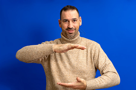 快乐的西班牙裔男子穿着棕色高领毛衣 双手之间展示着一些东西 产品营销广告概念与蓝色背景上的复制空间图片