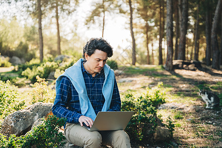 快乐开朗的人带着笔记本电脑坐在户外自然森林公园的岩石上 在山区度假时使用电脑打字 写博客 浏览的自由职业者 自由职业者 远程工作图片