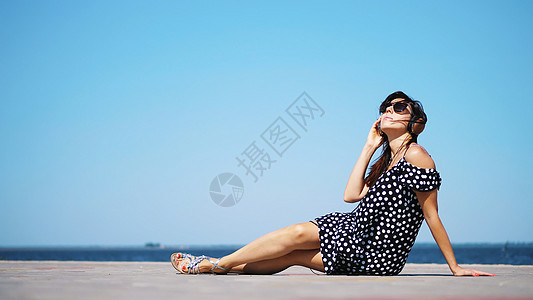 戴着太阳镜和太阳裙 戴着大耳机 在海滩上 在炎热的夏日 靠近海边的智能手机上听音乐的美丽黑发女孩旅行成人晴天青年幸福假期耳机音乐图片