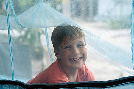 微笑的男孩用蚊子网在睡床上睡觉 童年白日梦快乐 看看 笑吧 肖像情绪化金发公园蓝色乐趣衬衫欢乐空气生活脚趾图片