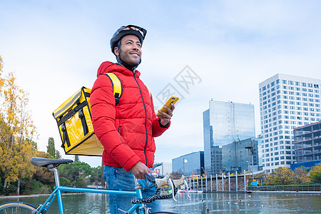 食品运送概念 在城市里 带着背背包智能手机微笑着的非裔美国人驾驶员命令外卖黑人邮递员运输自行车商业保温袋成人工作图片